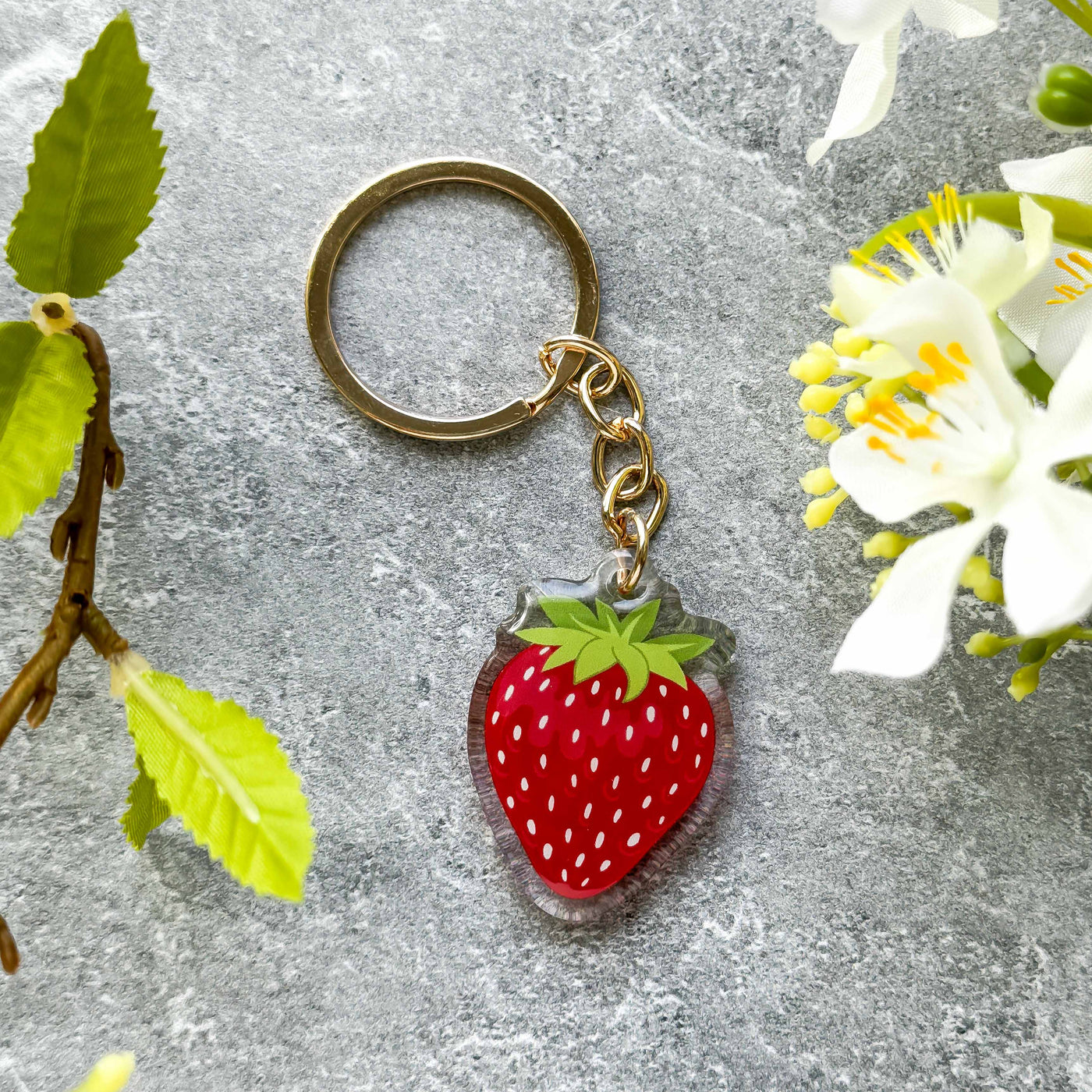 Strawberry-shaped keychain on a gray surface with green leaves and white flowers.