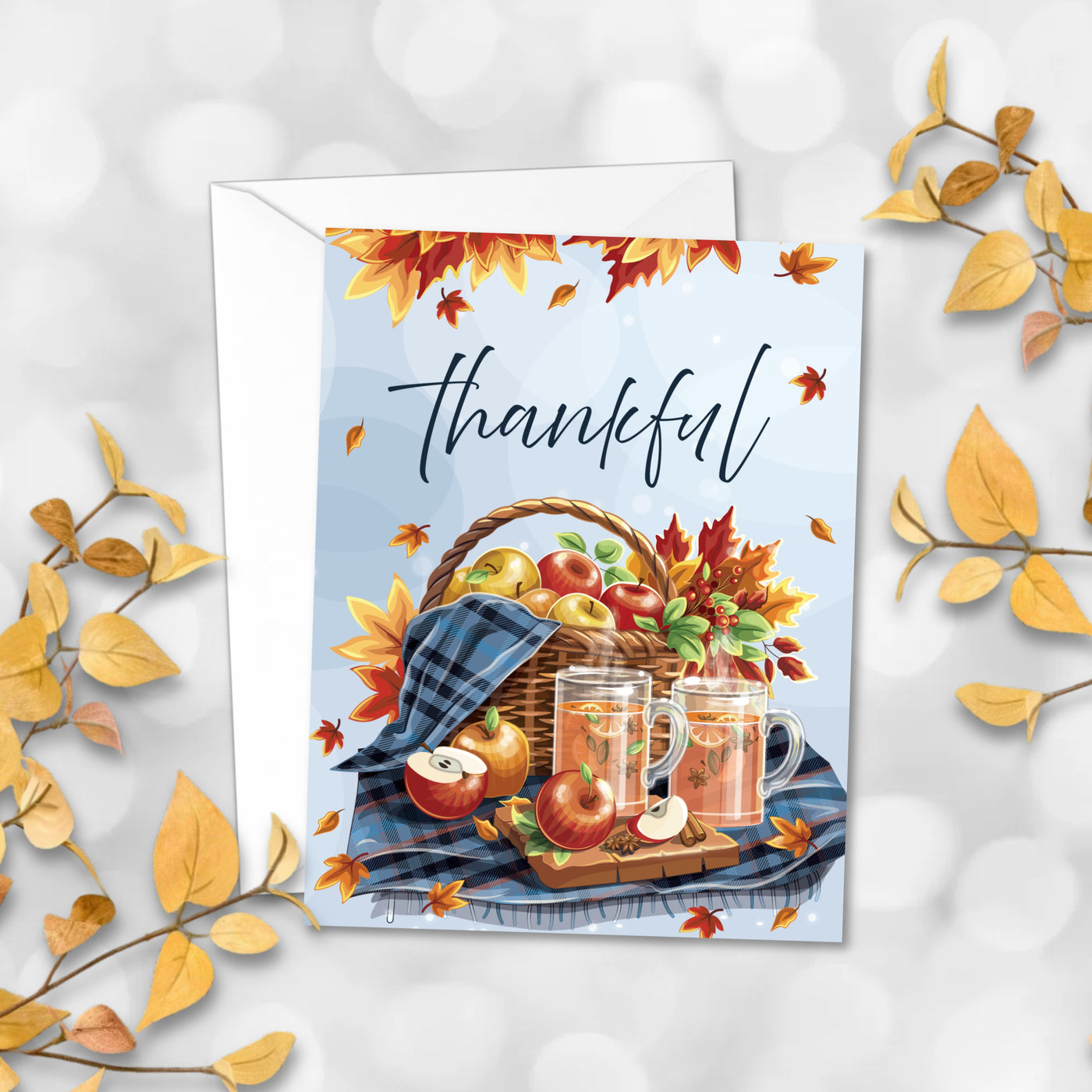 Greeting card with autumn theme, basket of apples and glasses of juice, surrounded by fall leaves on a light background.