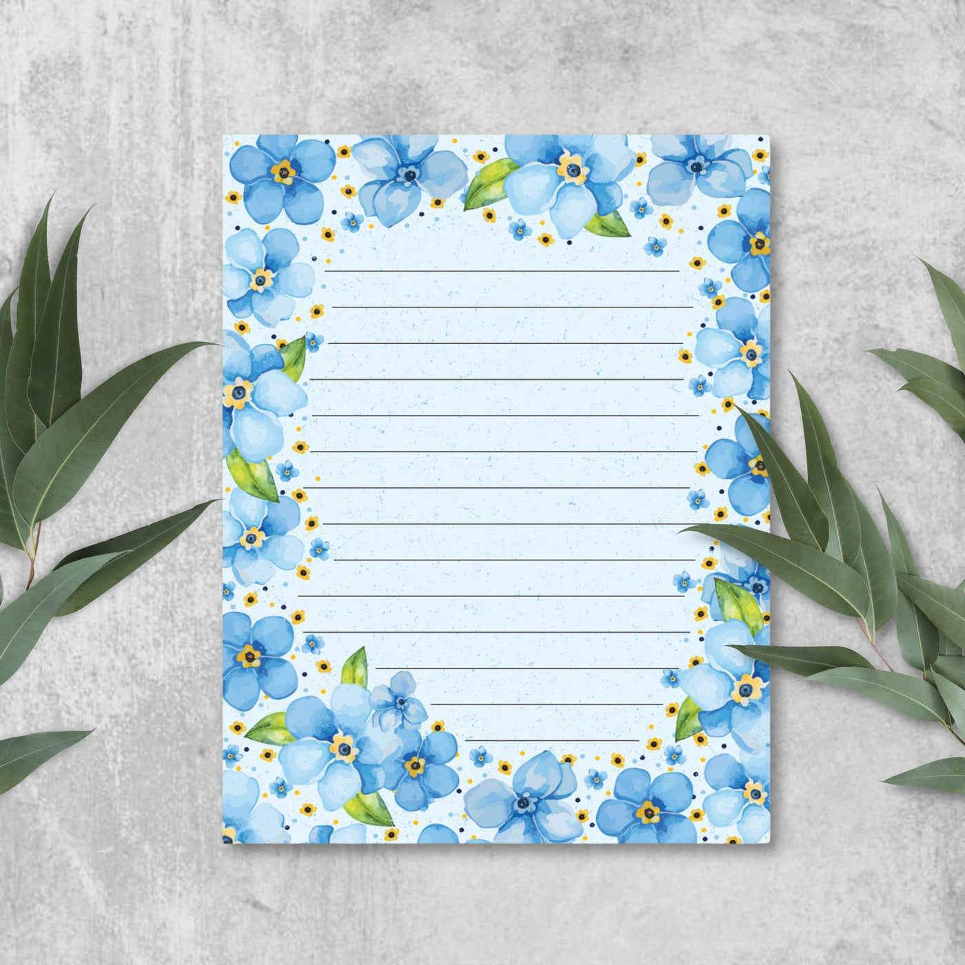 Floral-themed notepad with blue flowers on a light blue background, surrounded by green leaves on a gray surface.