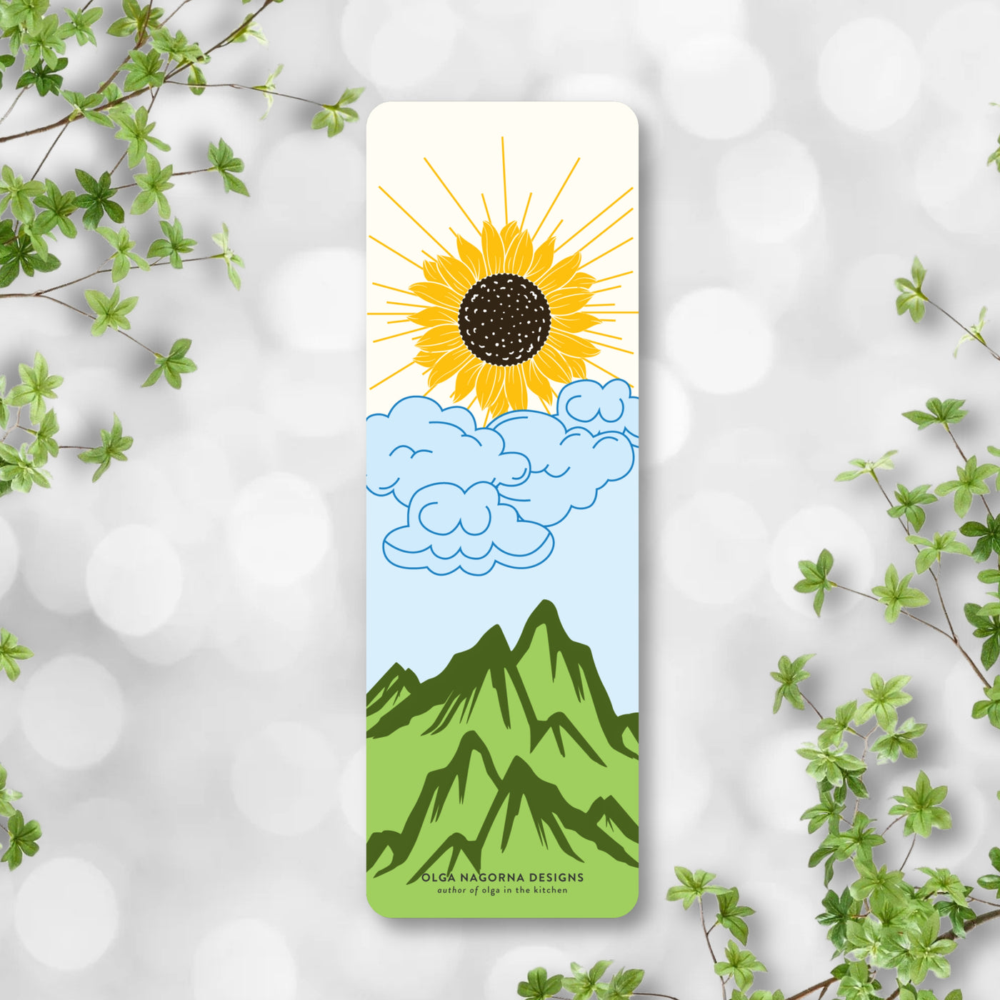 Bookmark with sun and mountain design on a light gray background with green leaves.