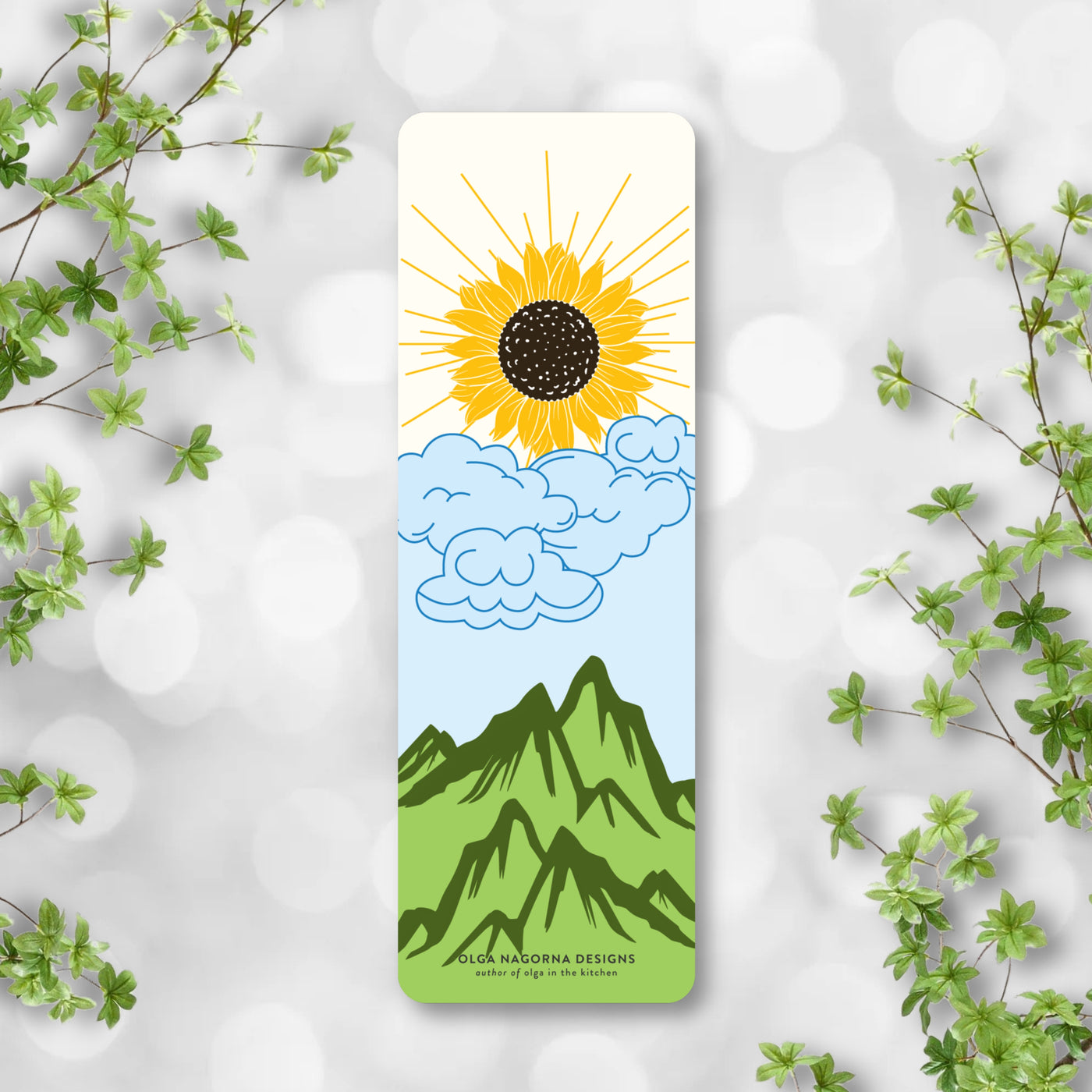 Bookmark with sun and mountain design on a light gray background with green leaves.